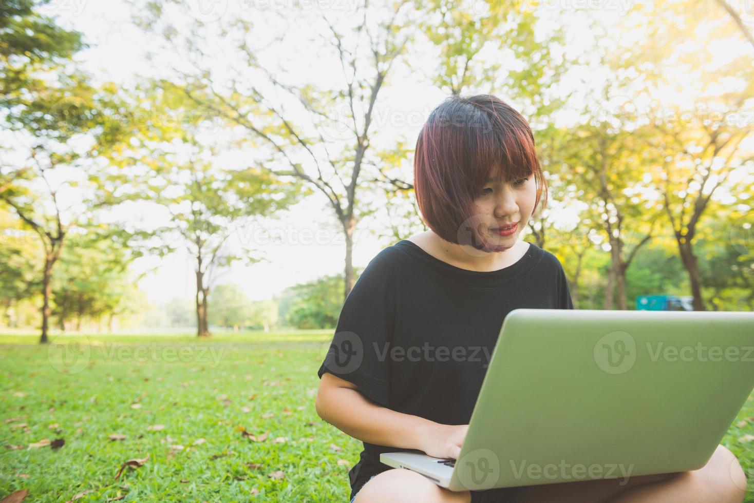 le gambe della giovane donna asiatica sull'erba verde con il computer portatile aperto. le mani della ragazza sulla tastiera. concetto di apprendimento a distanza. felice hipster giovane donna asiatica che lavora al computer portatile nel parco. studente che studia all'aperto. foto