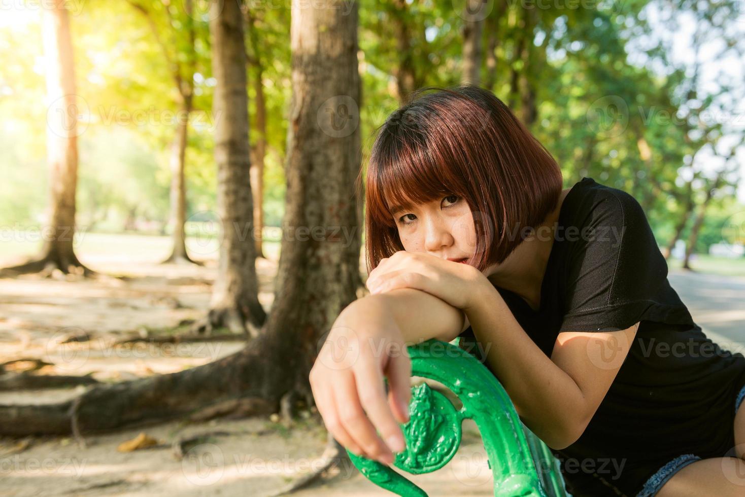 giovane donna asiatica seduta all'aperto sulla panchina pubblica e sdraiata lungo la panchina nel parco in una calda giornata di sole. la giovane donna rilassa la mente e il corpo nel parco. attività all'aperto nel concetto di parco. foto