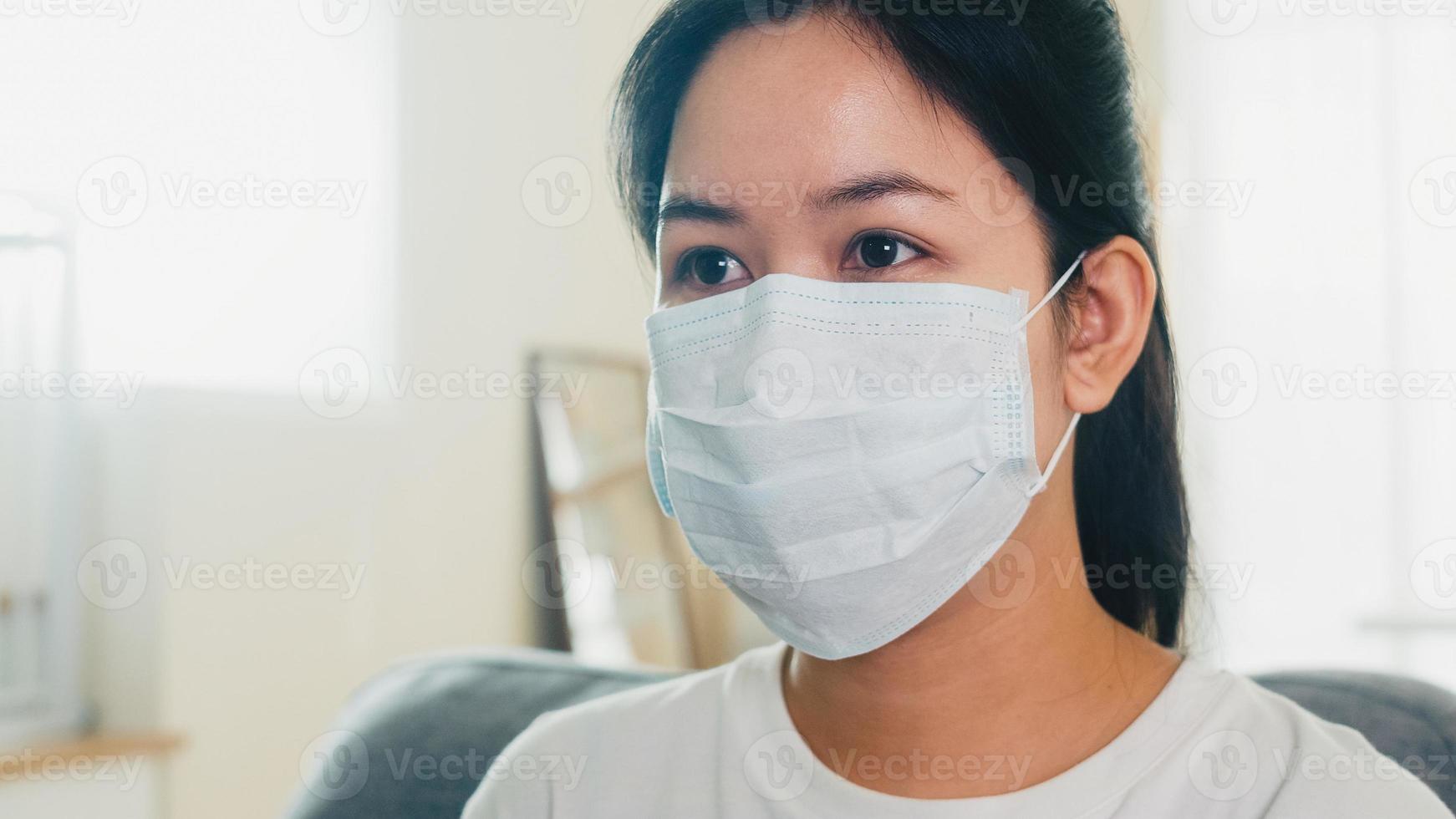 donna d'affari asiatica depressa che indossa una maschera protettiva seduta sul divano nel soggiorno di casa quando l'allontanamento sociale resta a casa e il tempo di autoquarantena, pandemia in Cina, concetto di coronavirus. foto