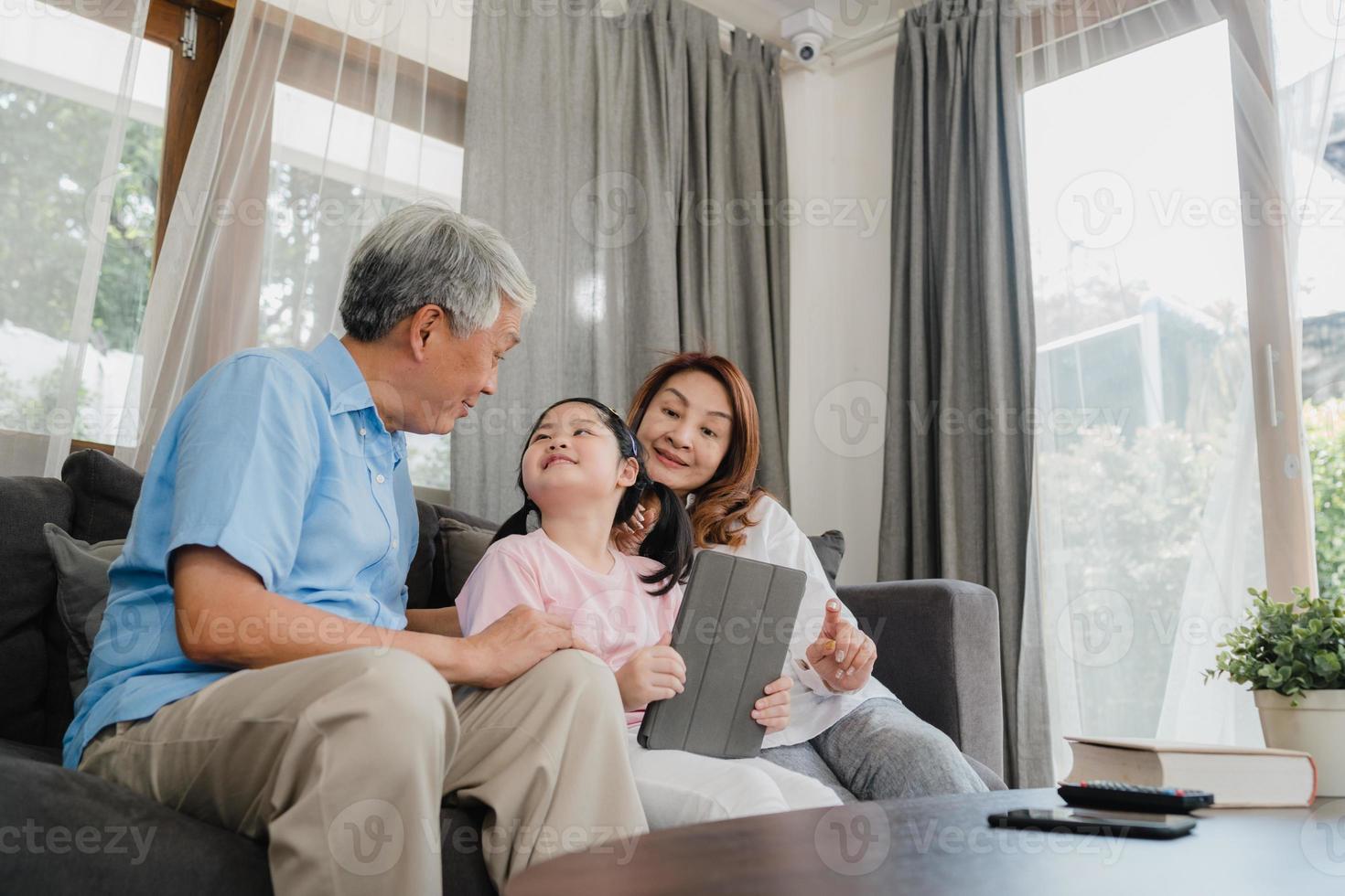 nonni asiatici e nipote utilizzando tablet a casa. cinese anziano, nonno e nonna felici trascorrono il tempo in famiglia rilassandosi con la ragazza che controlla i social media, sdraiati sul divano nel concetto del soggiorno foto