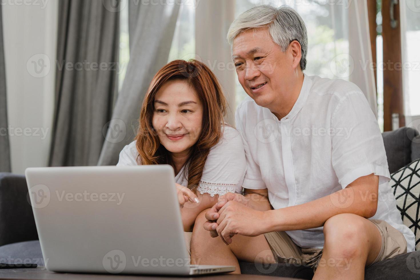 coppia senior asiatica che utilizza il computer portatile a casa. nonni cinesi anziani asiatici, navigare in internet per controllare i social media mentre si è sdraiati sul divano nel soggiorno a casa concetto. foto