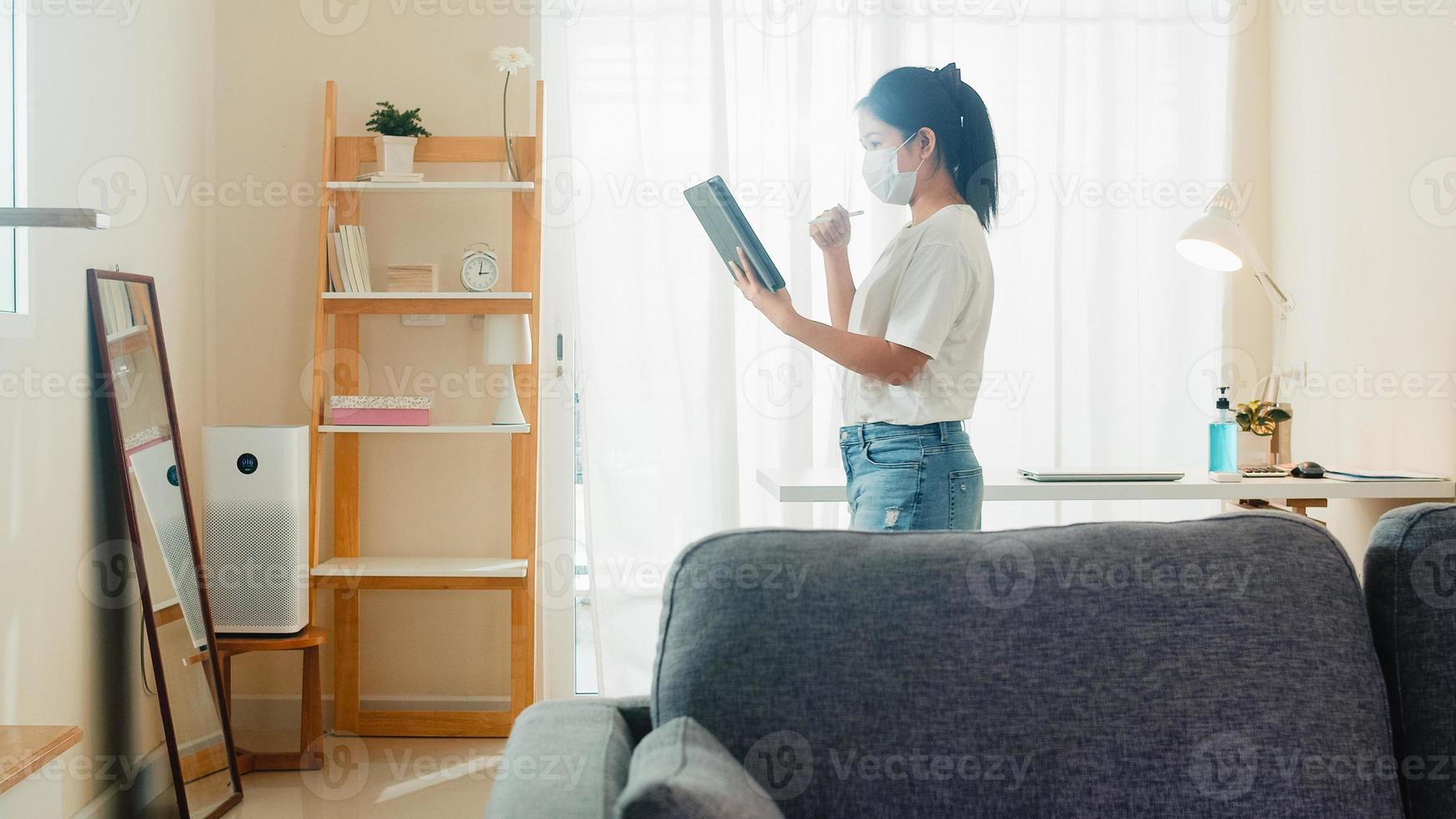 donna d'affari asiatica che indossa maschera utilizzando tablet videoconferenza chiamata di lavoro con il cliente nel soggiorno da casa quando l'allontanamento sociale rimane a casa e il tempo di autoquarantena, concetto di coronavirus. foto