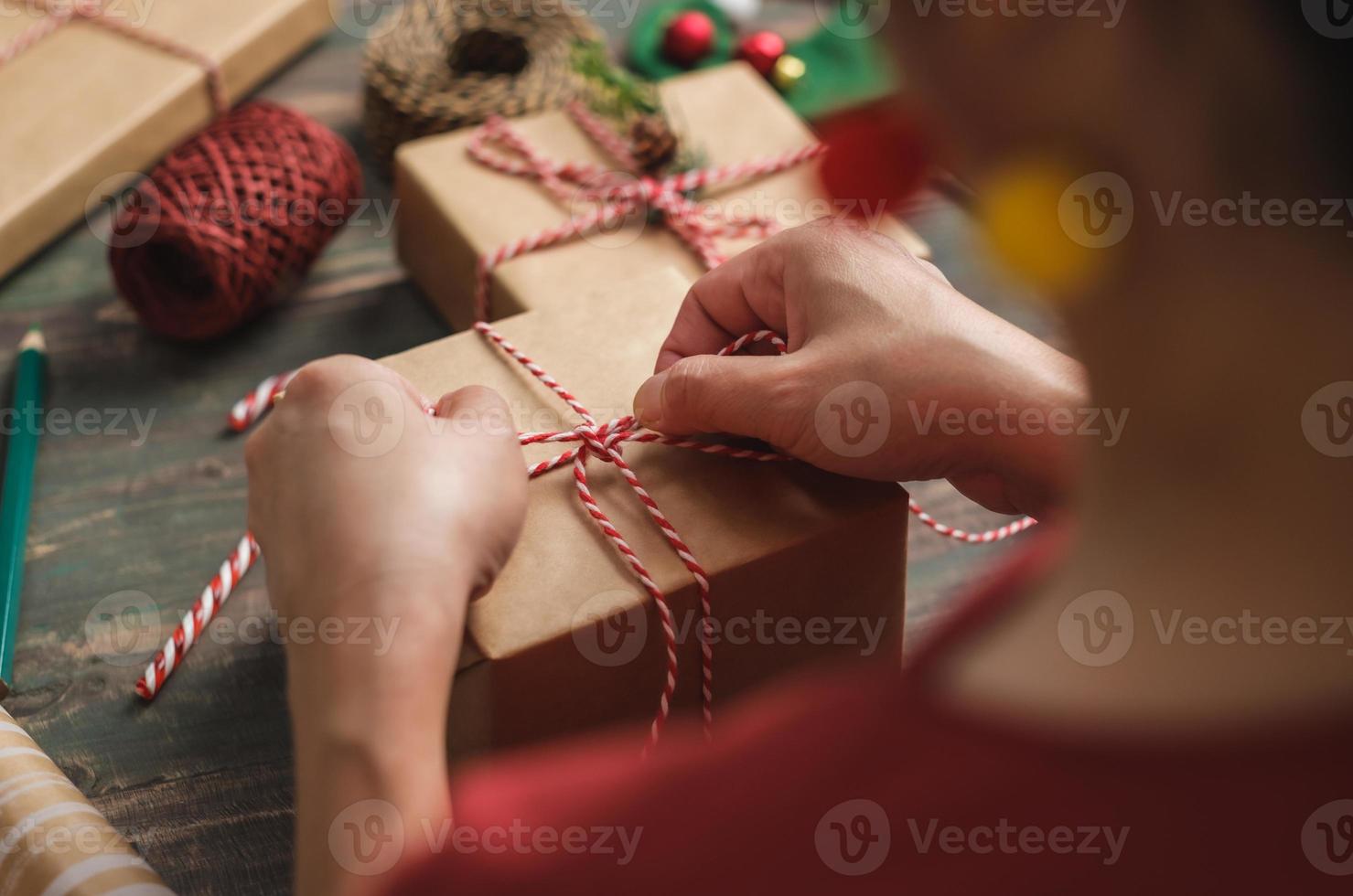 donna che fa una scatola regalo natalizia fatta a mano con carta marrone ordito con decorazioni natalizie su tavola di legno foto