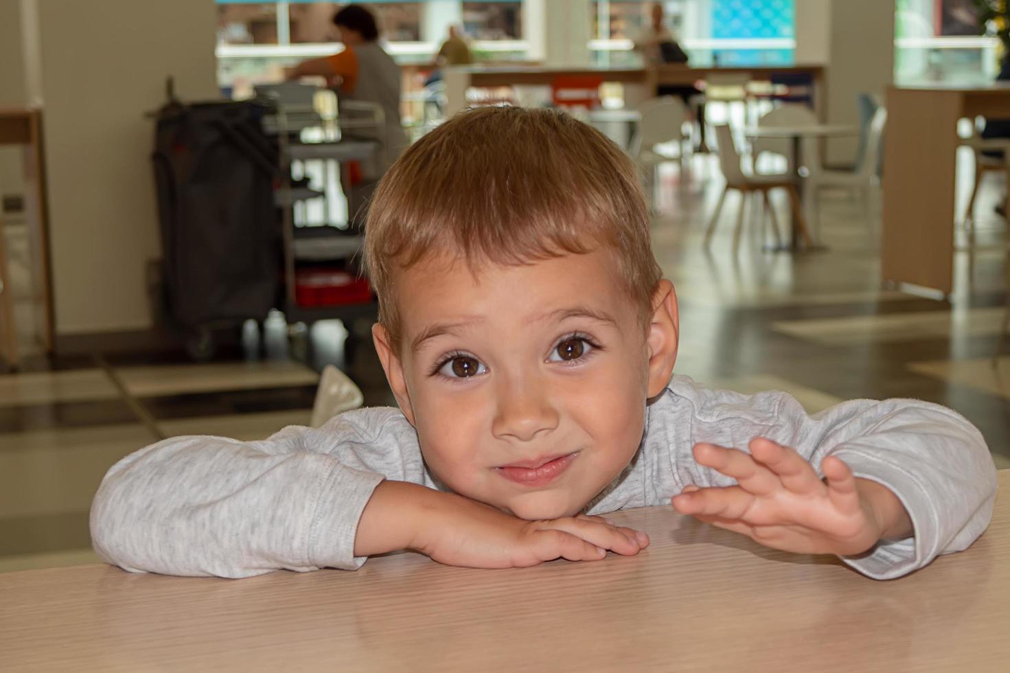 il ragazzino bello e allegro fa le smorfie in un caffè a un tavolo nella food court del centro commerciale. shopping. foto