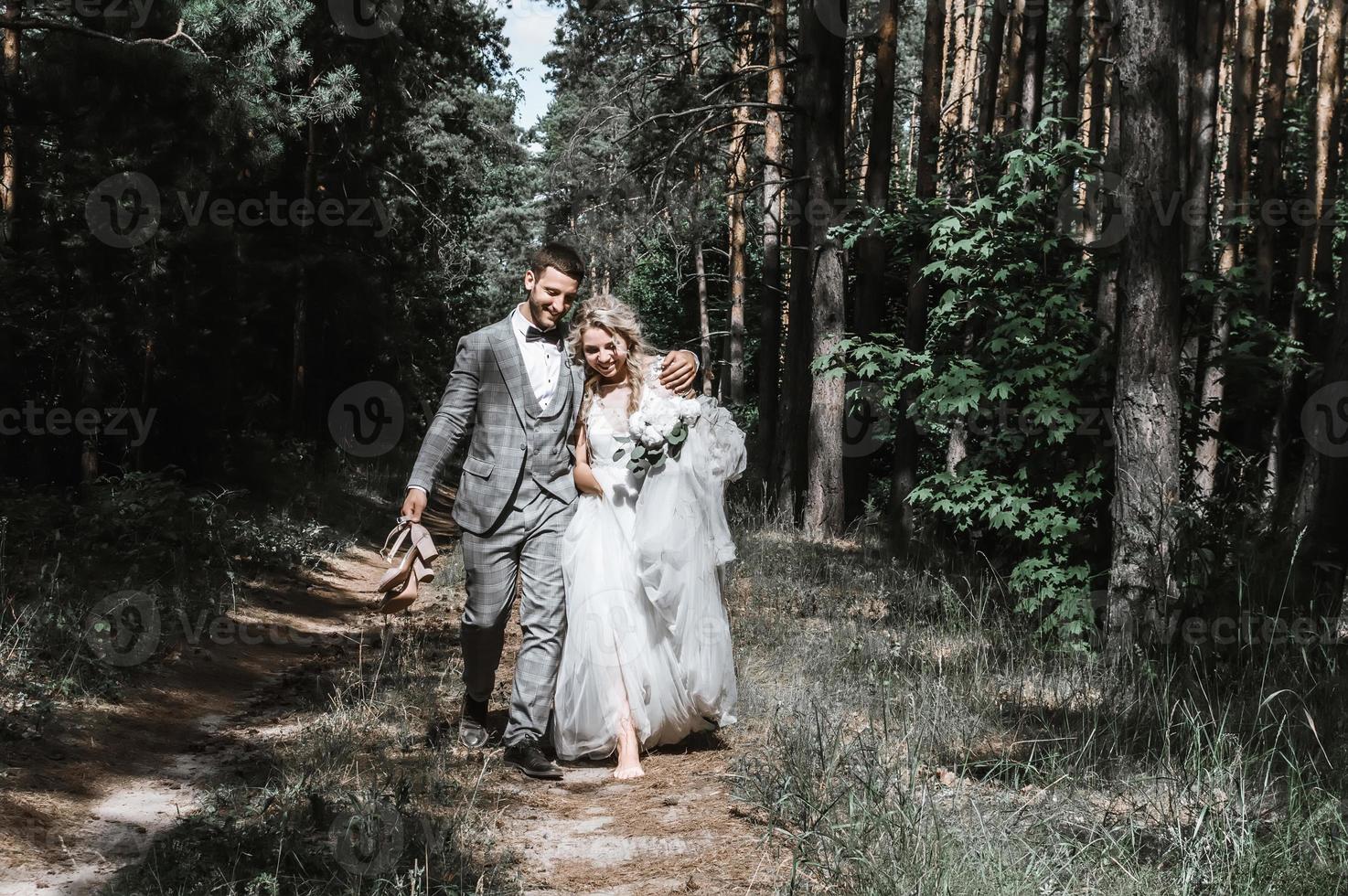 gli sposi attraversano il bosco abbracciati. sposa a piedi nudi, lo sposo porta le scarpe. giornata di sole estivo. foto