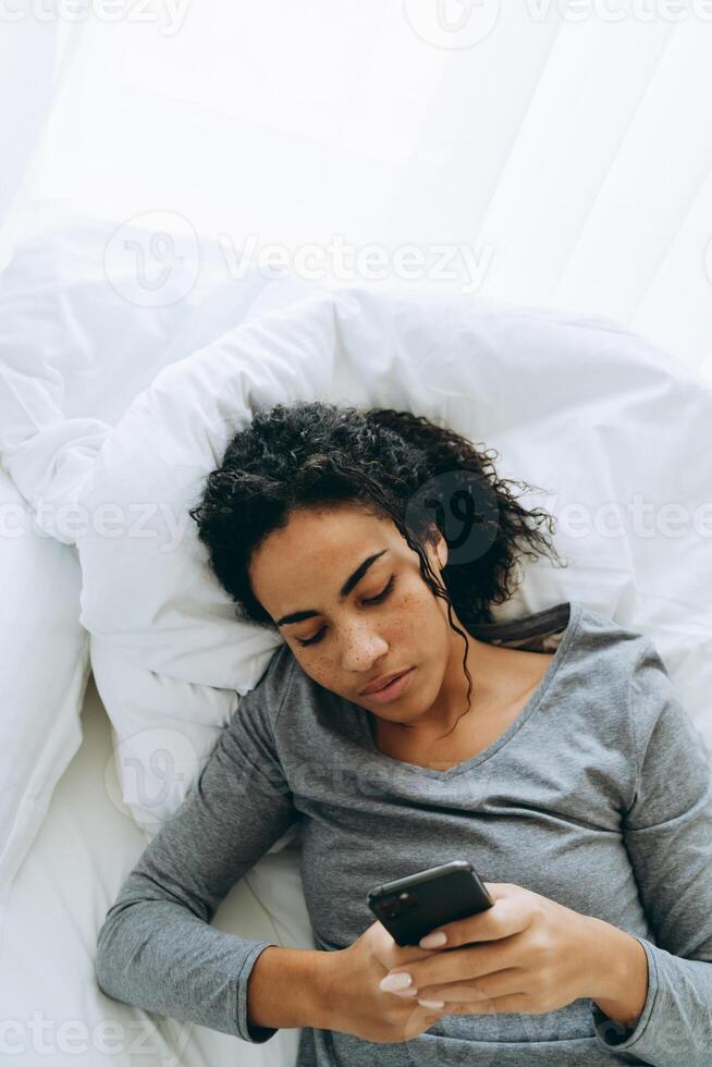 giovane donna di colore che usa il cellulare mentre giace a letto foto