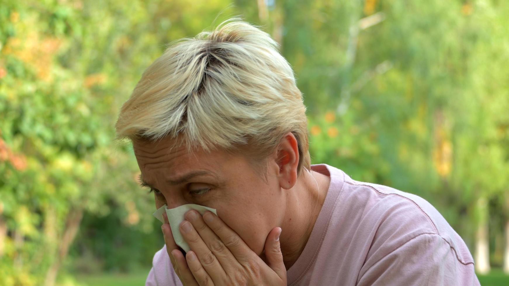 una ragazza con i capelli biondi e un taglio di capelli corto starnutisce per le allergie e si asciuga il naso con un tovagliolo sullo sfondo della natura verde foto