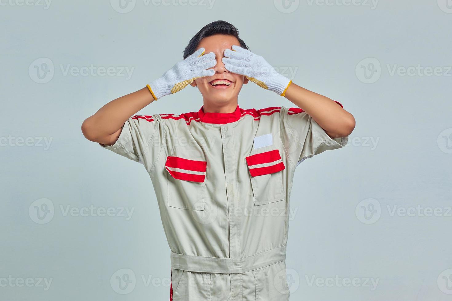 Sorridente bell'uomo che indossa l'uniforme del meccanico che copre gli occhi con le mani su sfondo grigio foto