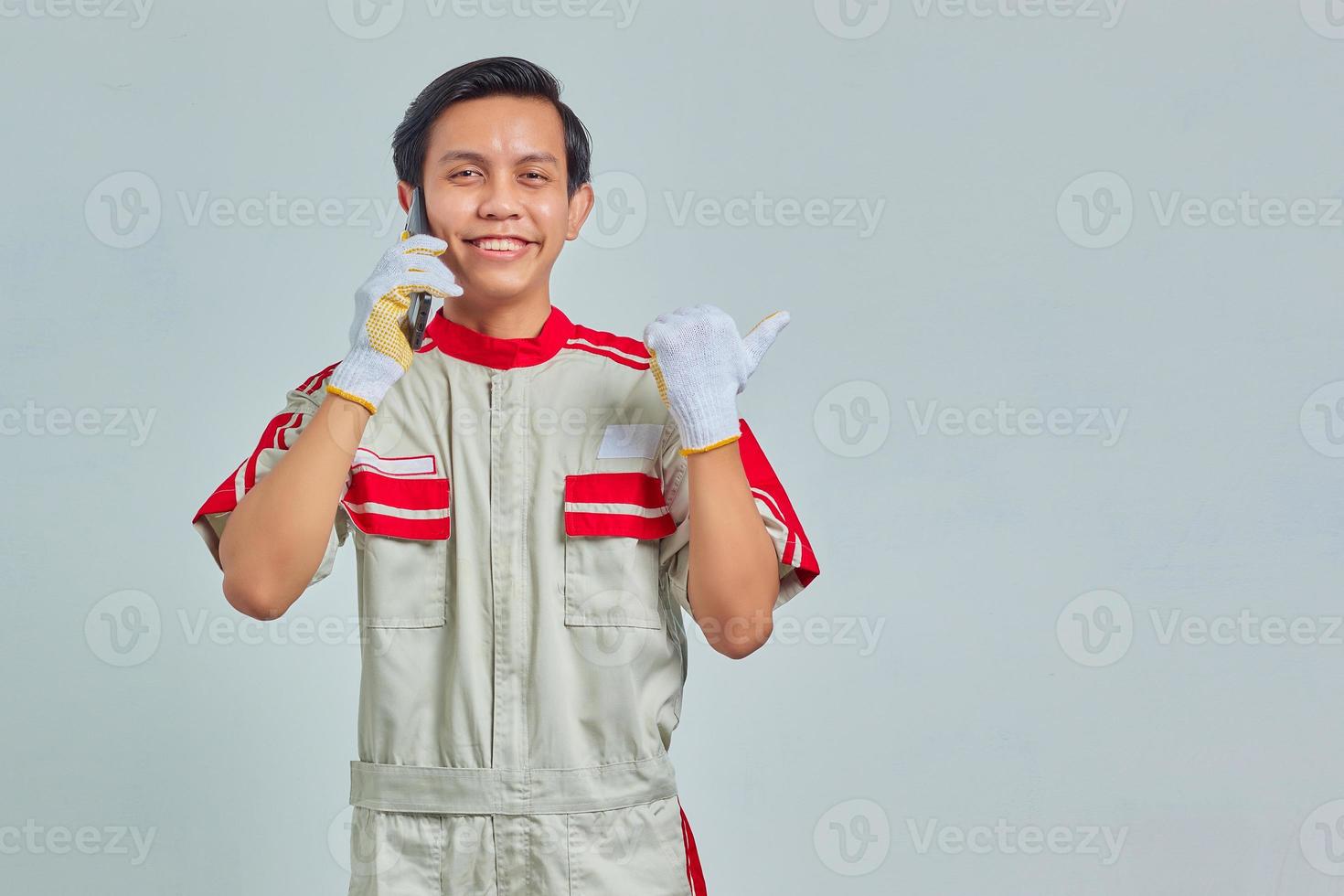 ritratto di felice giovane bello meccanico uomo che indossa l'uniforme parlando al cellulare e puntando lateralmente su sfondo grigio foto