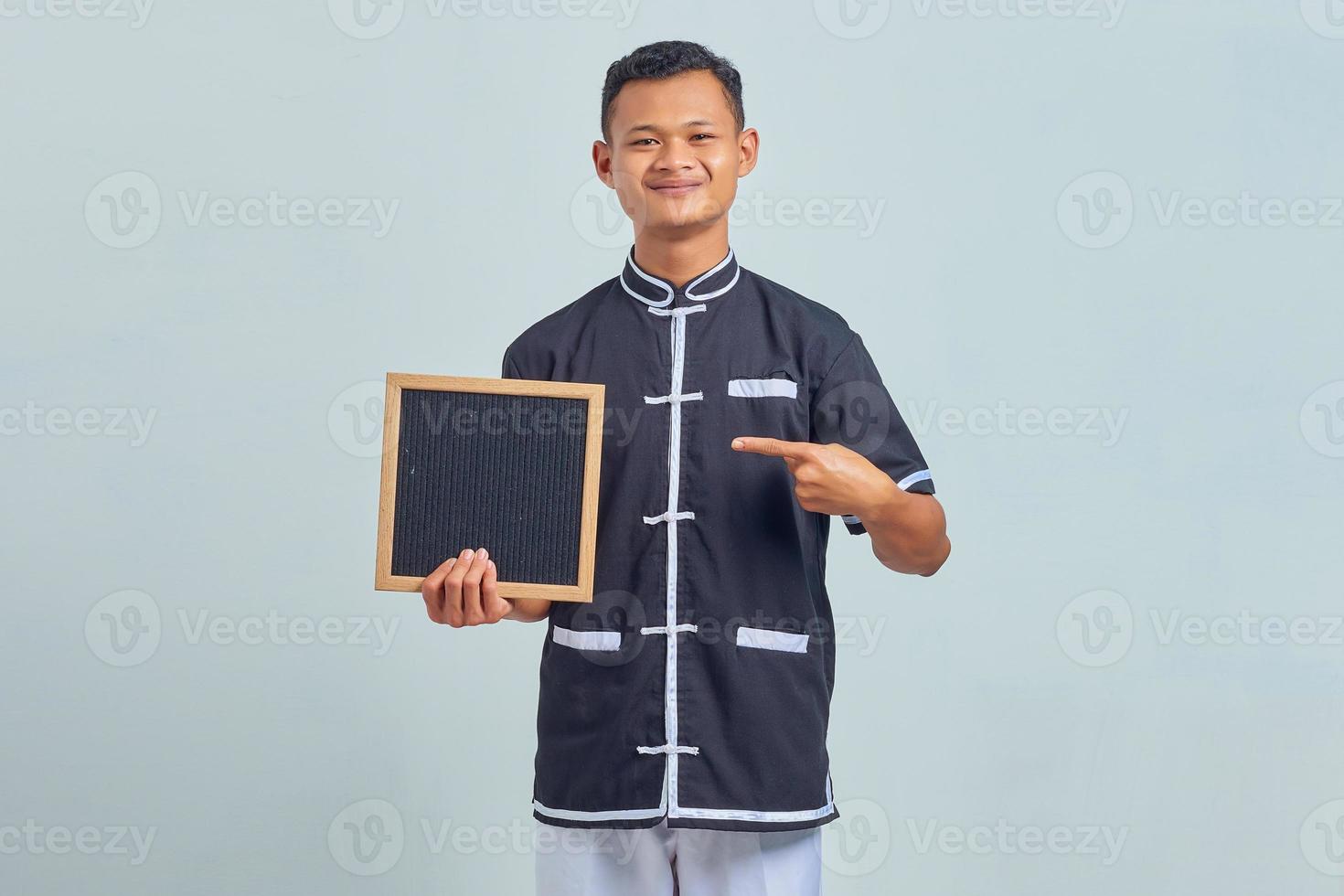 ritratto di allegro giovane asiatico che indossa l'uniforme di karate che punta al bordo bianco con il dito su sfondo grigio foto