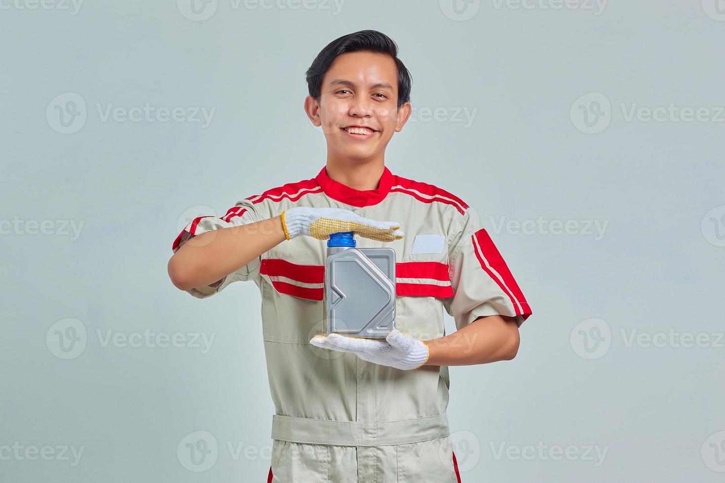 Ritratto di un bell'uomo sorridente che indossa l'uniforme del meccanico che mostra la bottiglia di plastica dell'olio motore su sfondo grigio foto