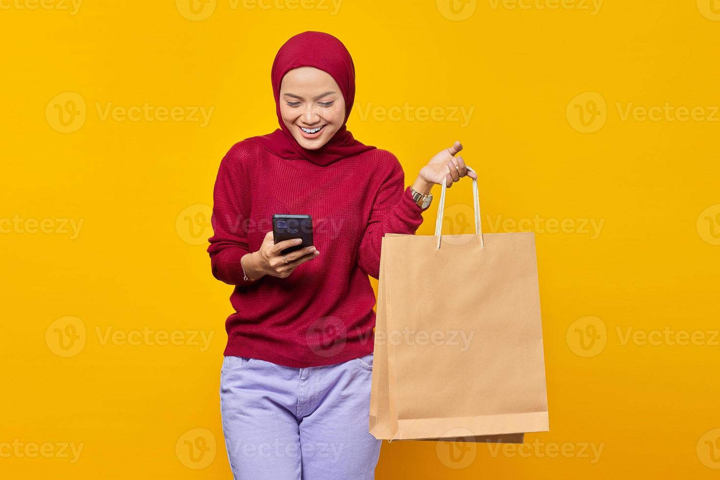 sorridente giovane donna asiatica che legge il messaggio sullo smartphone e mostra le borse della spesa su sfondo giallo foto