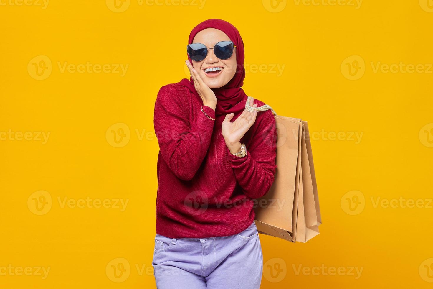 felice giovane donna asiatica con la mano sulle guance e tenendo le borse della spesa su sfondo giallo foto