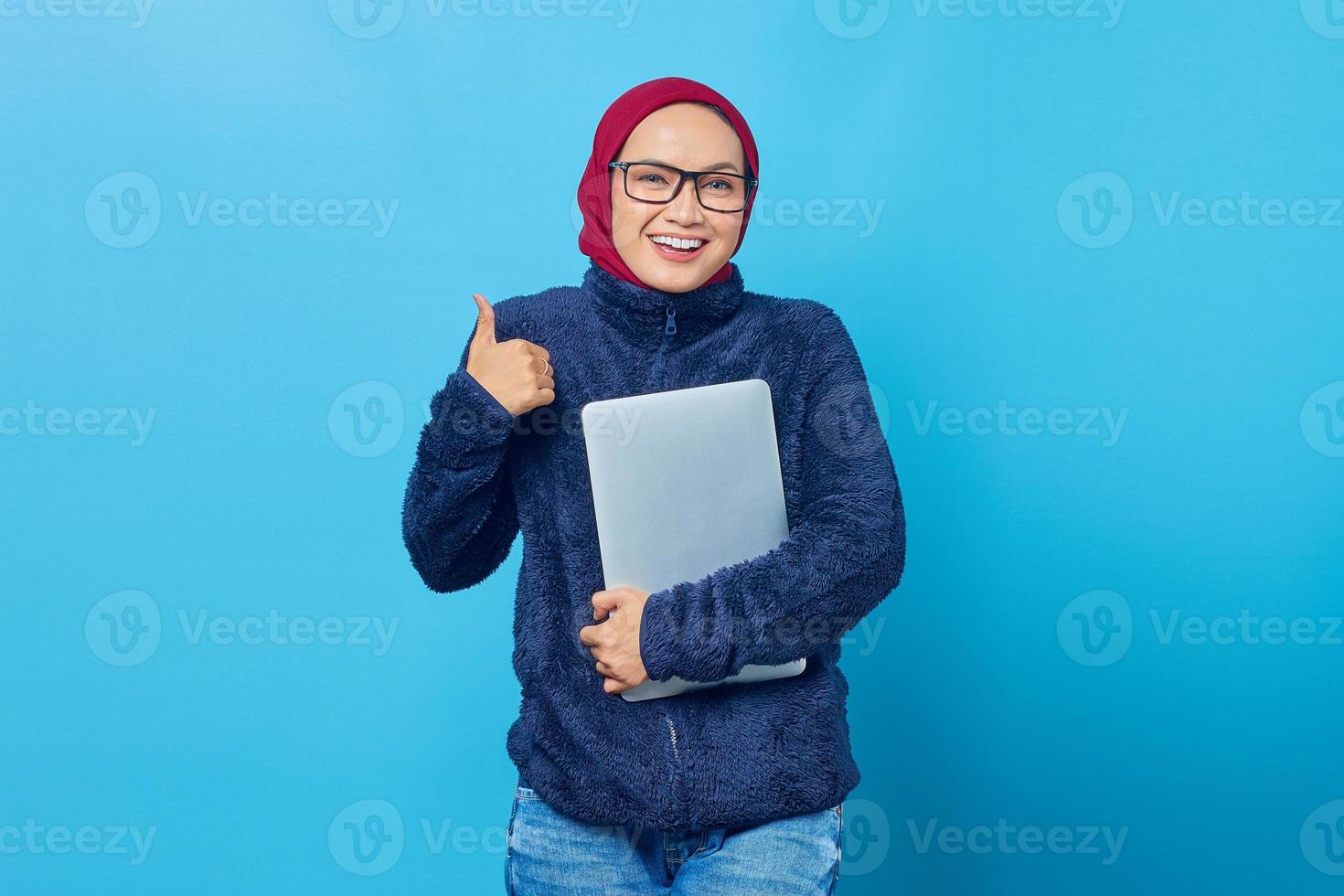 allegro e bellissimo giovane studente che indossa una giacca blu che tiene il computer portatile e dà i pollici in su su sfondo blu foto