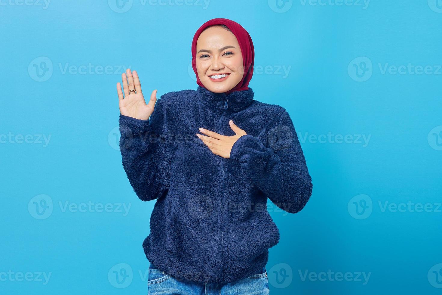 ritratto di giovane donna asiatica sorridente che giura con le mani sul petto e con i palmi aperti su sfondo blu foto