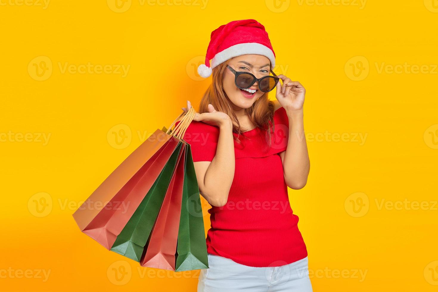 Sorridente giovane donna asiatica in cappello di Babbo Natale e occhiali da sole che tengono le borse della spesa isolate su sfondo giallo foto