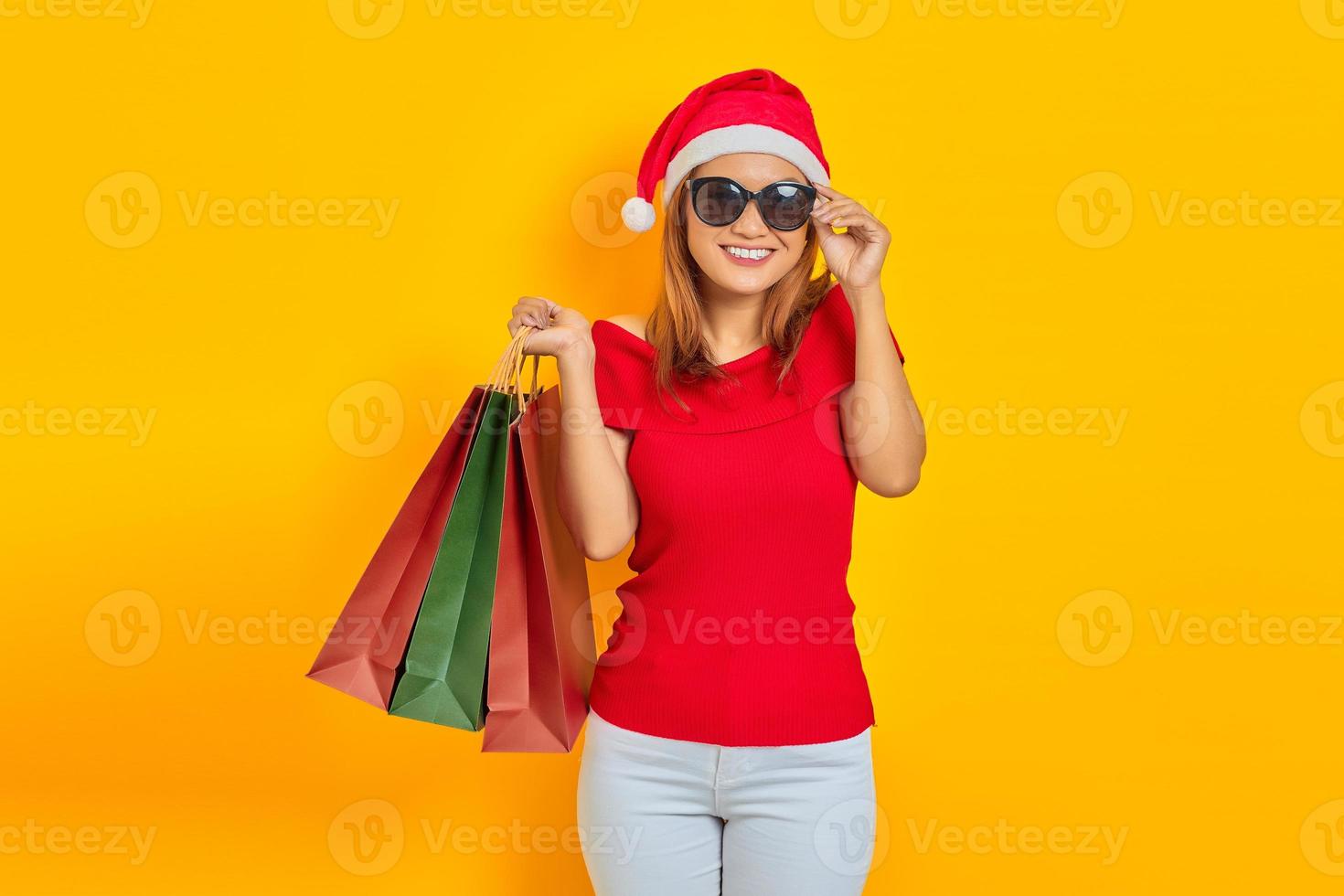 Allegra giovane donna asiatica in cappello di Babbo Natale e occhiali da sole che tengono le borse della spesa isolate su sfondo giallo foto