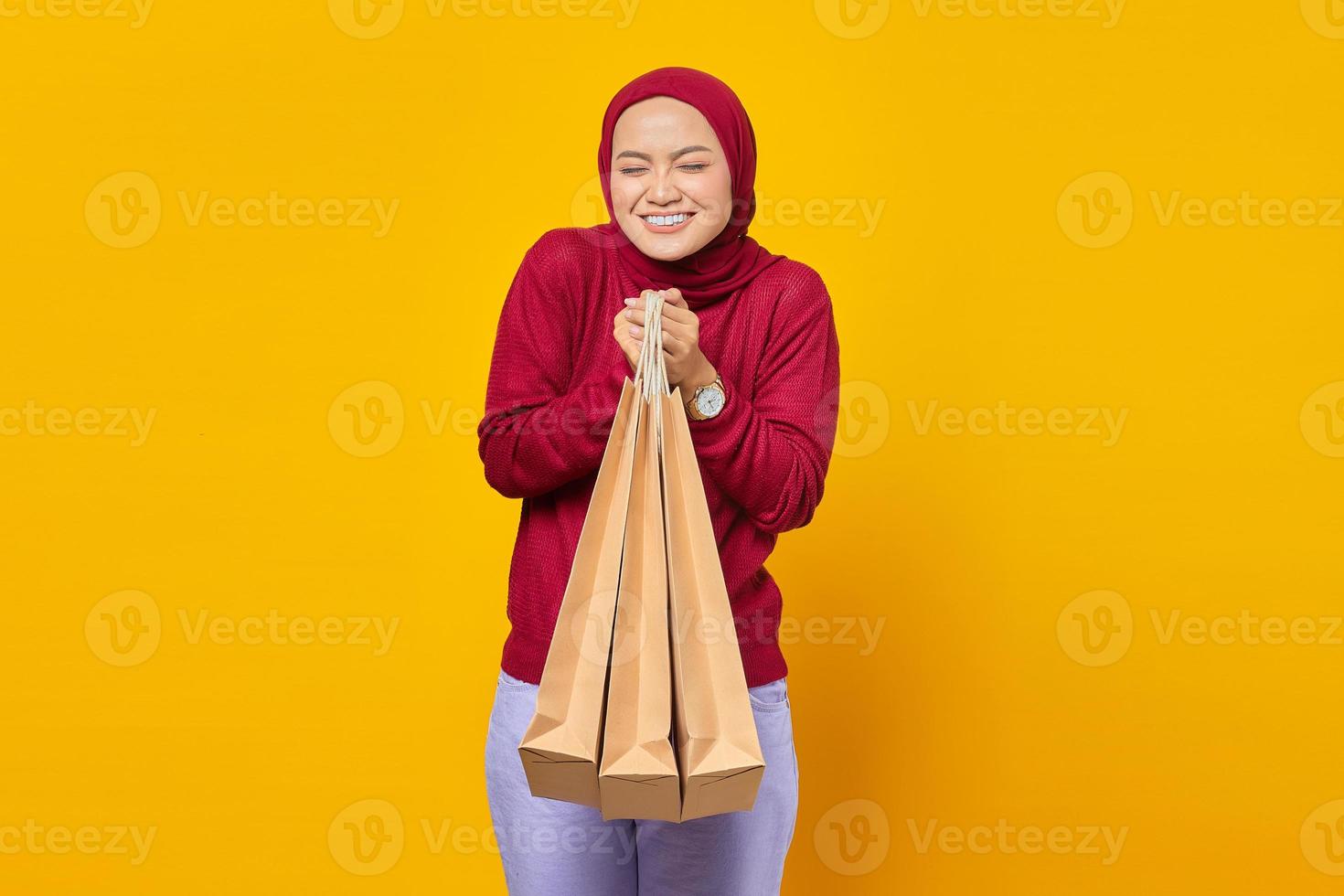 donna asiatica eccitata e sorridente che chiude gli occhi e tiene in mano le borse della spesa su sfondo giallo foto