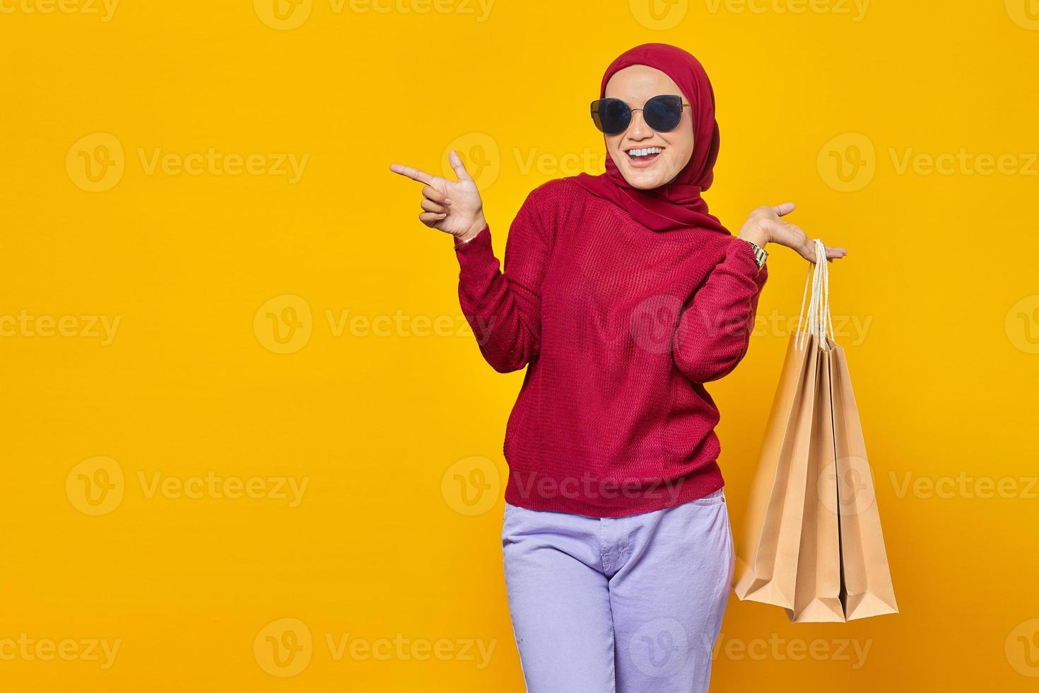 Allegra giovane donna asiatica che tiene le borse della spesa e punta il dito contro lo spazio della copia isolato su sfondo giallo foto