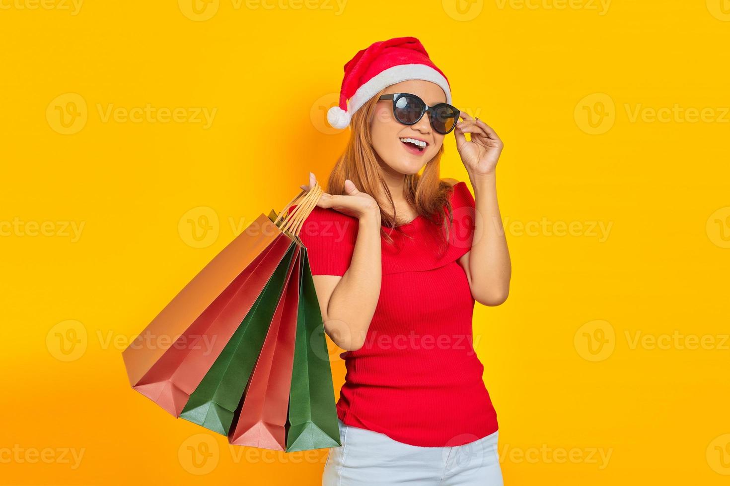 Sorridente giovane donna asiatica in cappello di Babbo Natale e occhiali da sole che tengono le borse della spesa isolate su sfondo giallo foto