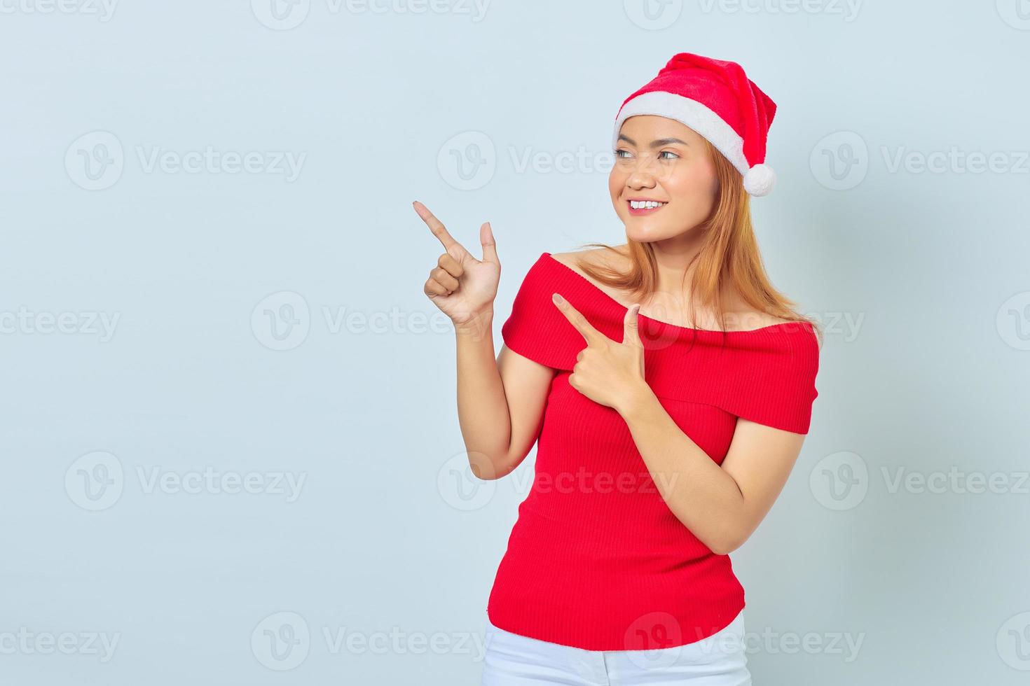 Sorridente giovane donna asiatica che indossa un abito rosso puntare il dito contro lo spazio della copia isolato su sfondo bianco foto