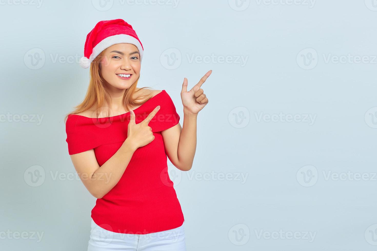 Sorridente giovane donna asiatica che indossa un abito rosso puntare il dito contro lo spazio della copia isolato su sfondo bianco foto