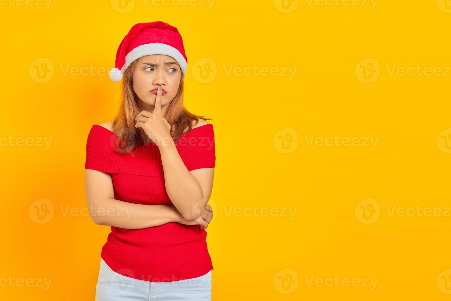 attraente giovane donna asiatica che indossa il cappello di Natale e fa il gesto di silenzio su sfondo giallo foto