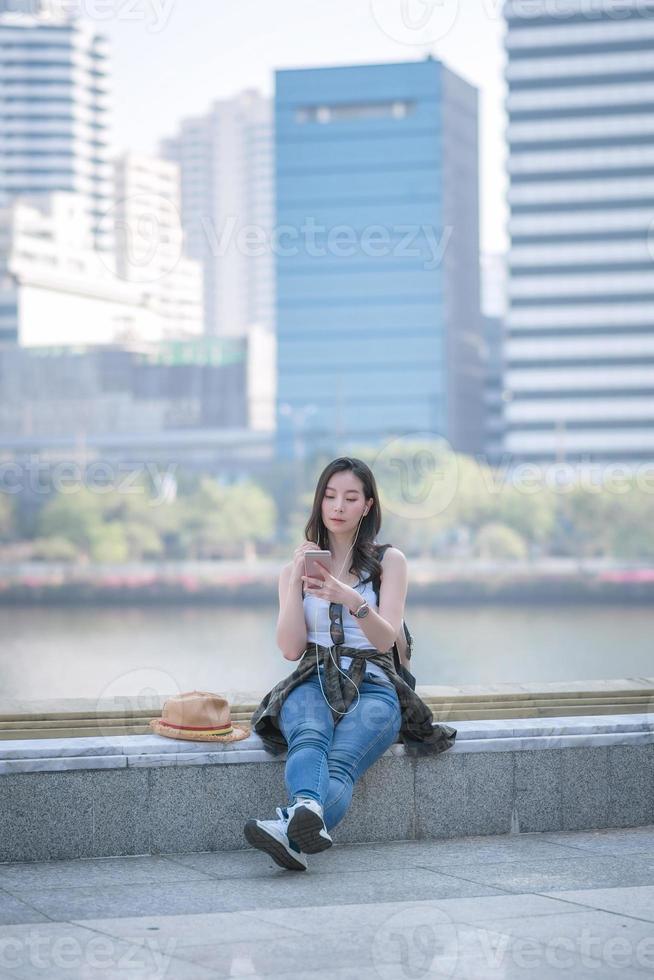 bella donna turistica asiatica che si rilassa e si diverte ad ascoltare la musica su uno smartphone nel centro urbano della città. viaggio di vacanza in estate. foto