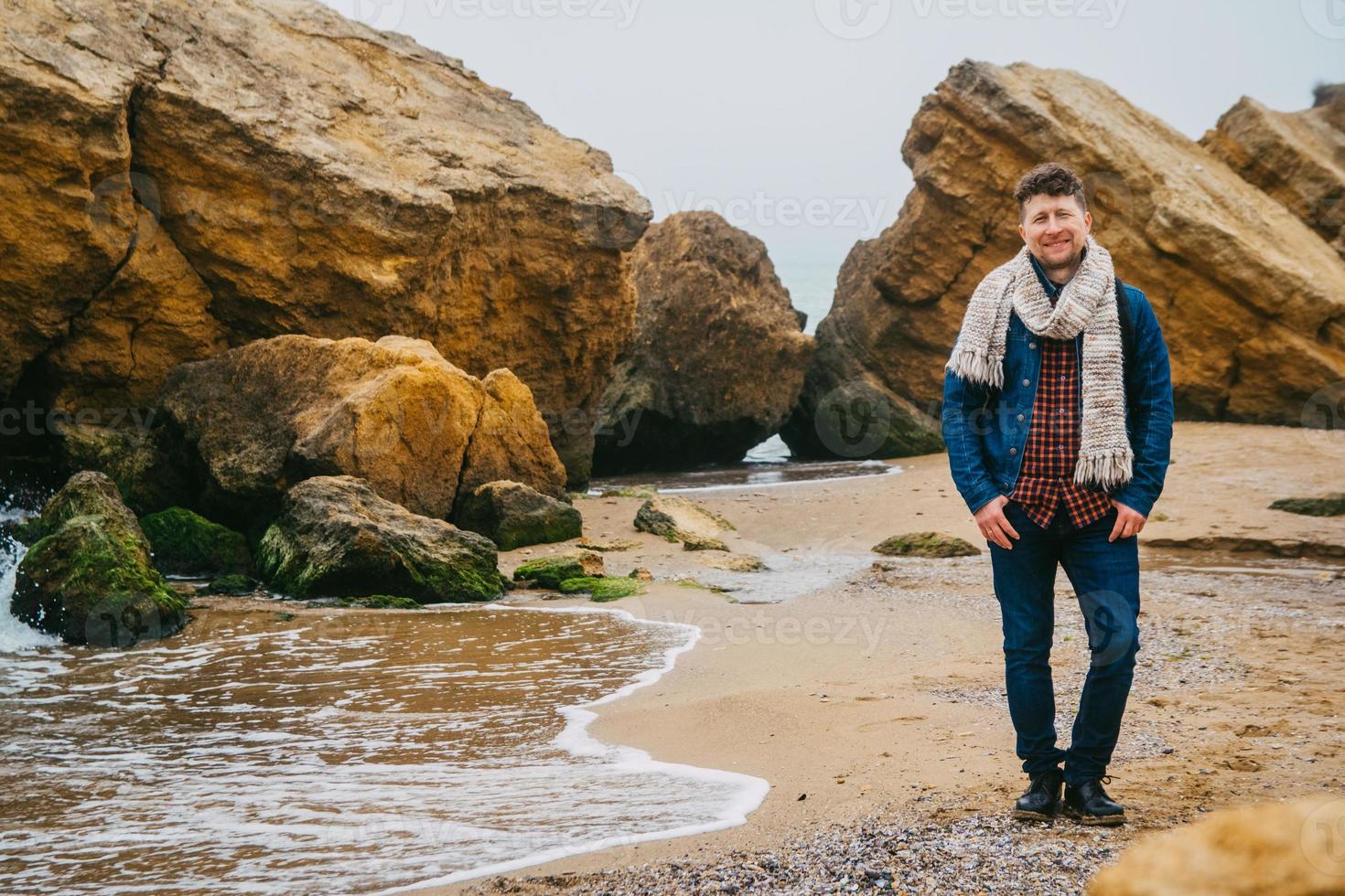 uomo viaggiatore con zaino in piedi sulla spiaggia sabbiosa in mezzo alle rocce sullo sfondo del mare foto