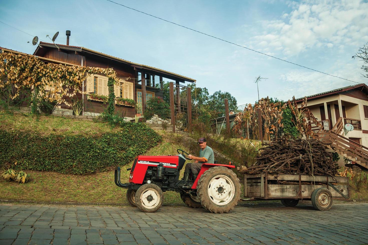 bento goncalves, brasile - 13 luglio 2019. trattore che tira il carrello con sottobosco su una strada lastricata in pietra, accanto a una casa di legno vicino a bento goncalves. un accogliente paese di campagna famoso per la sua produzione di vino. foto