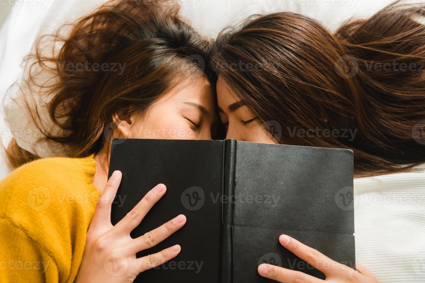 vista dall'alto di belle giovani donne asiatiche lesbica coppia felice bacia e sorride mentre si trova insieme a letto sotto il libro a casa. donne divertenti dopo il risveglio. coppia lesbica insieme al chiuso concetto. foto