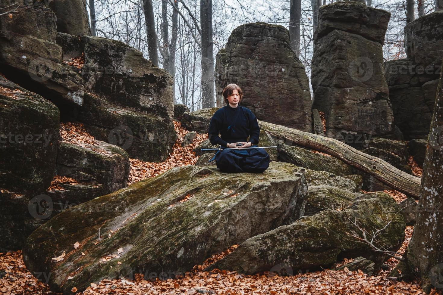 l'uomo in kimono nero con una spada medita e si concentra su rocce e sullo sfondo della foresta foto