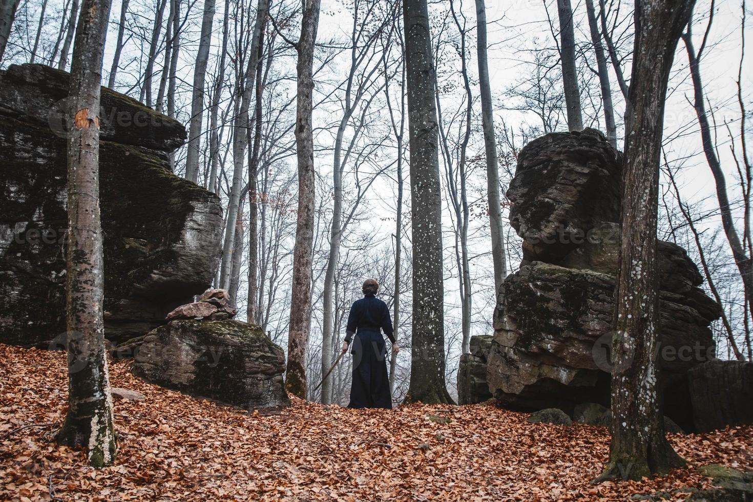 uomo in kimono nero che pratica arti marziali con una spada su rocce e sullo sfondo della foresta foto