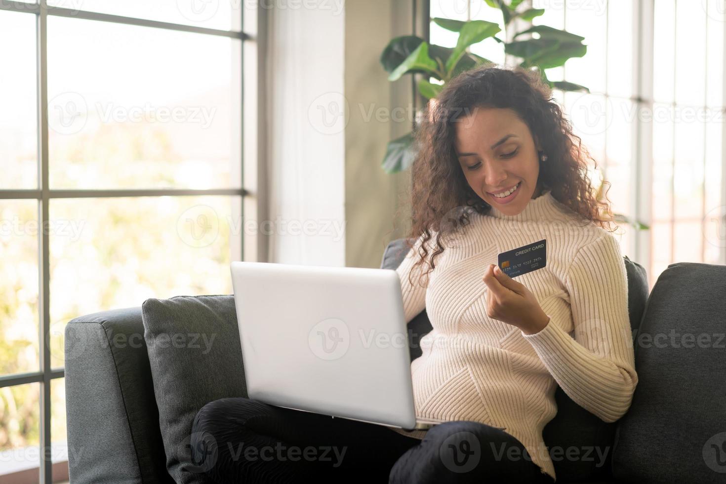 donna latina che utilizza laptop e mano che tiene la carta di credito per lo shopping sul divano foto