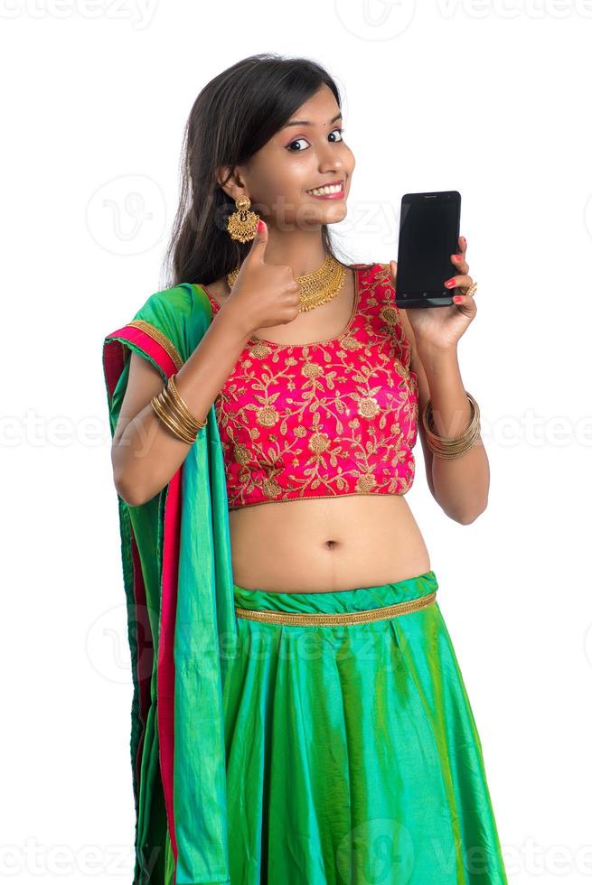 giovane ragazza indiana tradizionale che utilizza un telefono cellulare o uno smartphone e mostra uno smartphone con schermo vuoto su sfondo bianco foto