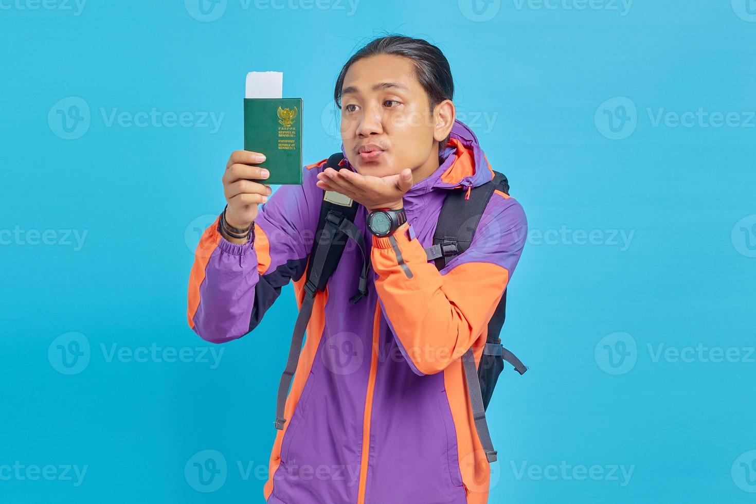 ritratto di giovane uomo asiatico che indossa una giacca viola che invia un bacio aereo al libro del passaporto isolato su sfondo blu foto