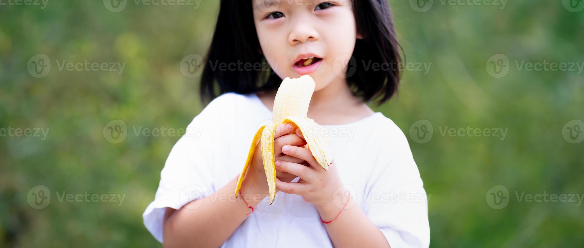 bambino felice di 4 anni mangia frutta matura che è dolce e deliziosa. bambino asiatico tiene la banana in mano. donna che indossa una maglietta bianca. foto