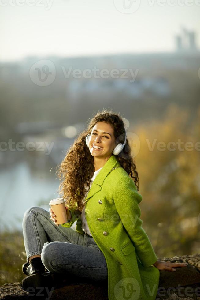 bella giovane donna che ascolta musica con smartphone e caffè da asporto in riva al fiume in una soleggiata giornata autunnale foto