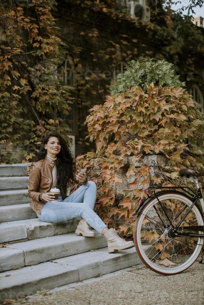 giovane donna con telefono cellulare bere caffè per andare alle scale in bicicletta il giorno d'autunno foto