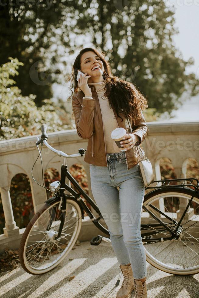giovane donna con telefono cellulare bere caffè da asporto in bicicletta il giorno d'autunno foto