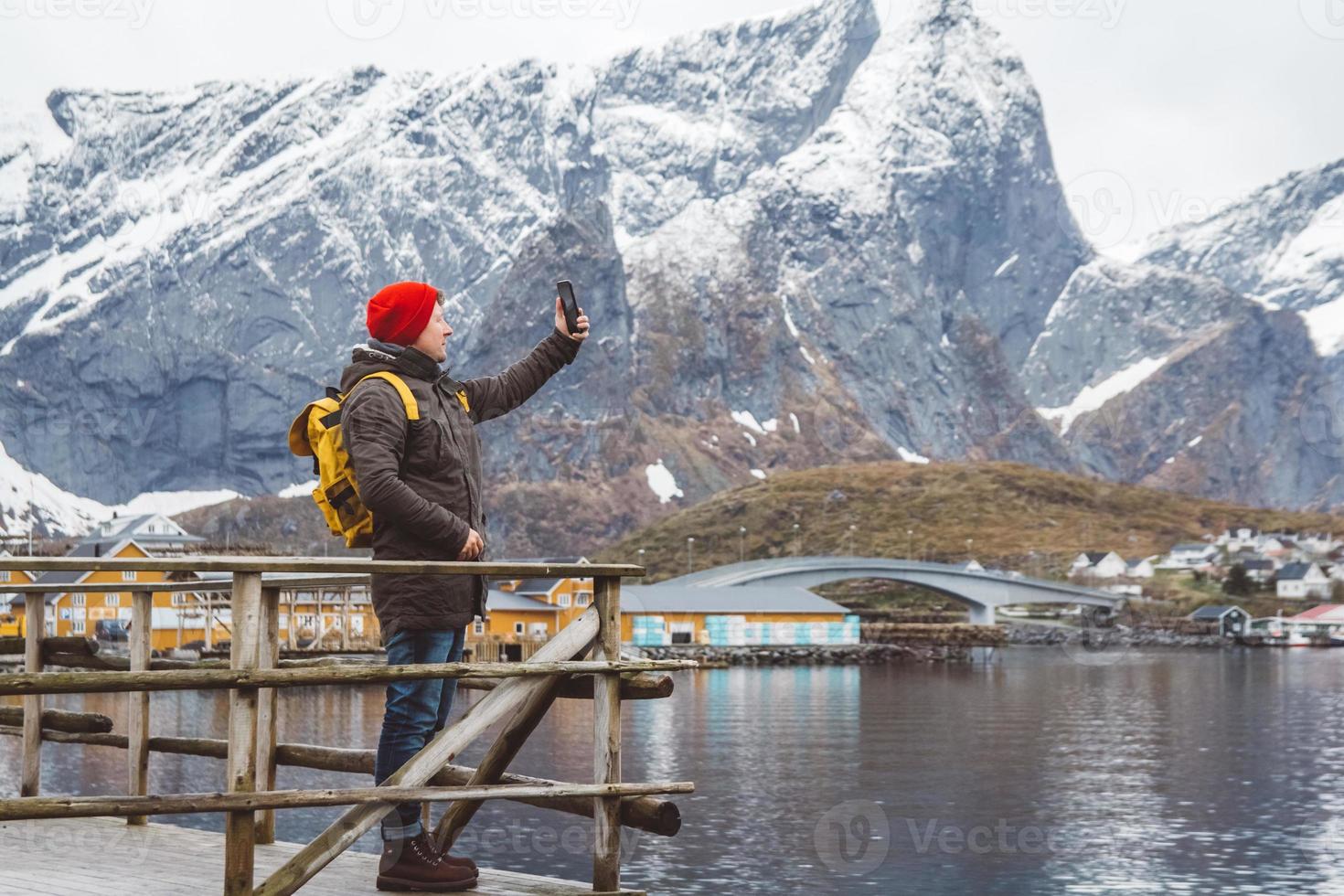 uomo viaggiatore che prende autoritratto con uno smartphone sullo sfondo di montagne innevate, rocce e laghi in piedi su un molo di legno. posto per testo o pubblicità foto