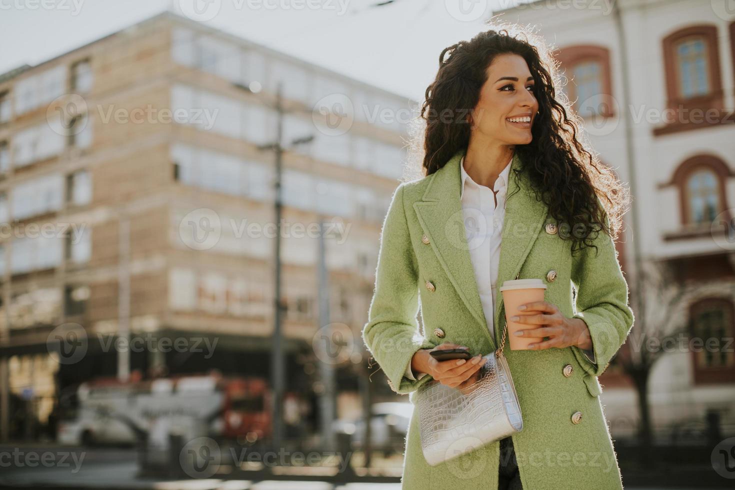 giovane donna che utilizza smartphone per strada e tiene caffè da asporto foto