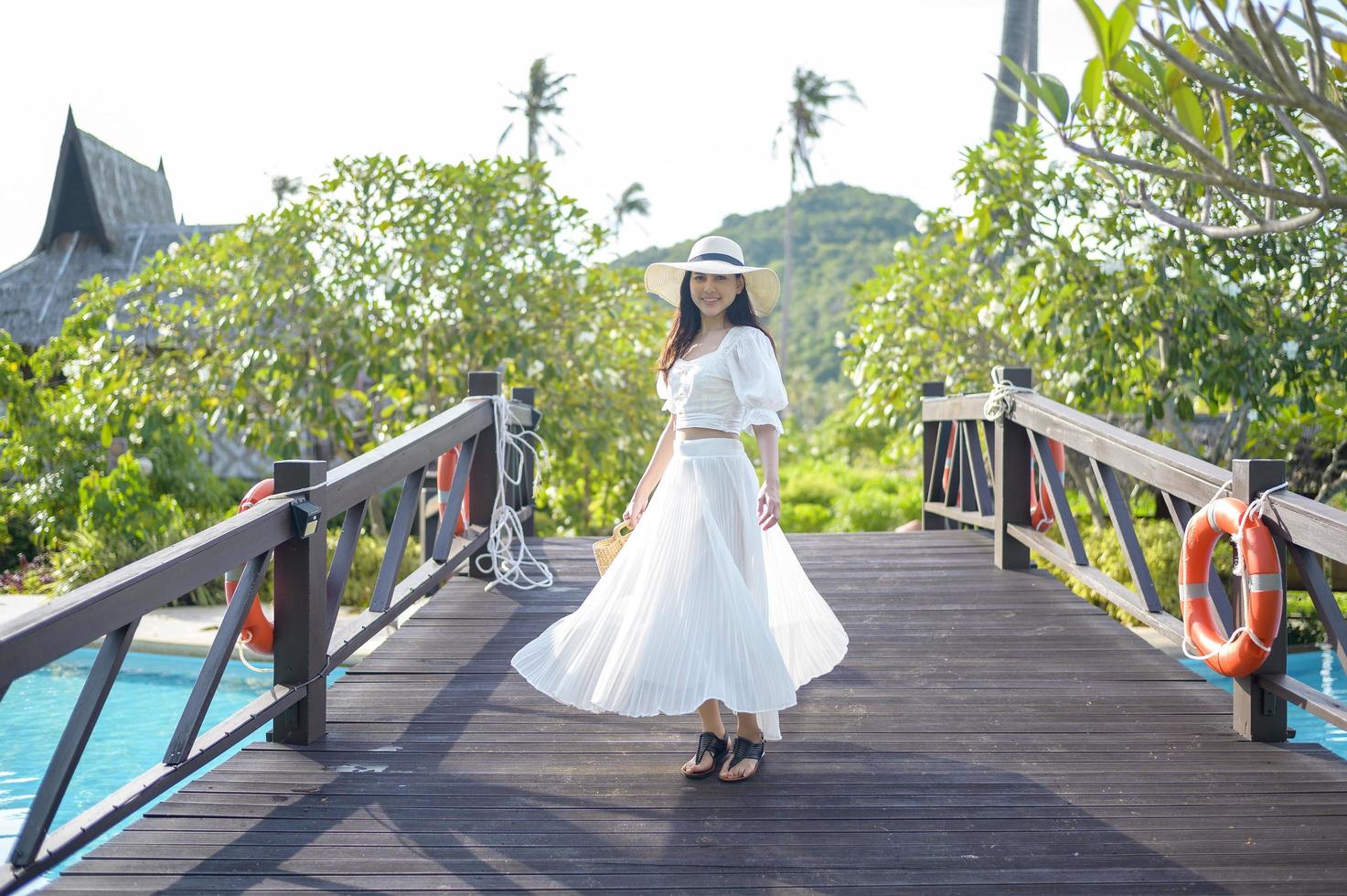 una bella donna felice in abito bianco godendo e in piedi sul ponte di legno sopra la piscina in un accogliente bungalow con giardino tropicale verde sull'isola di phi phi, tailandia foto