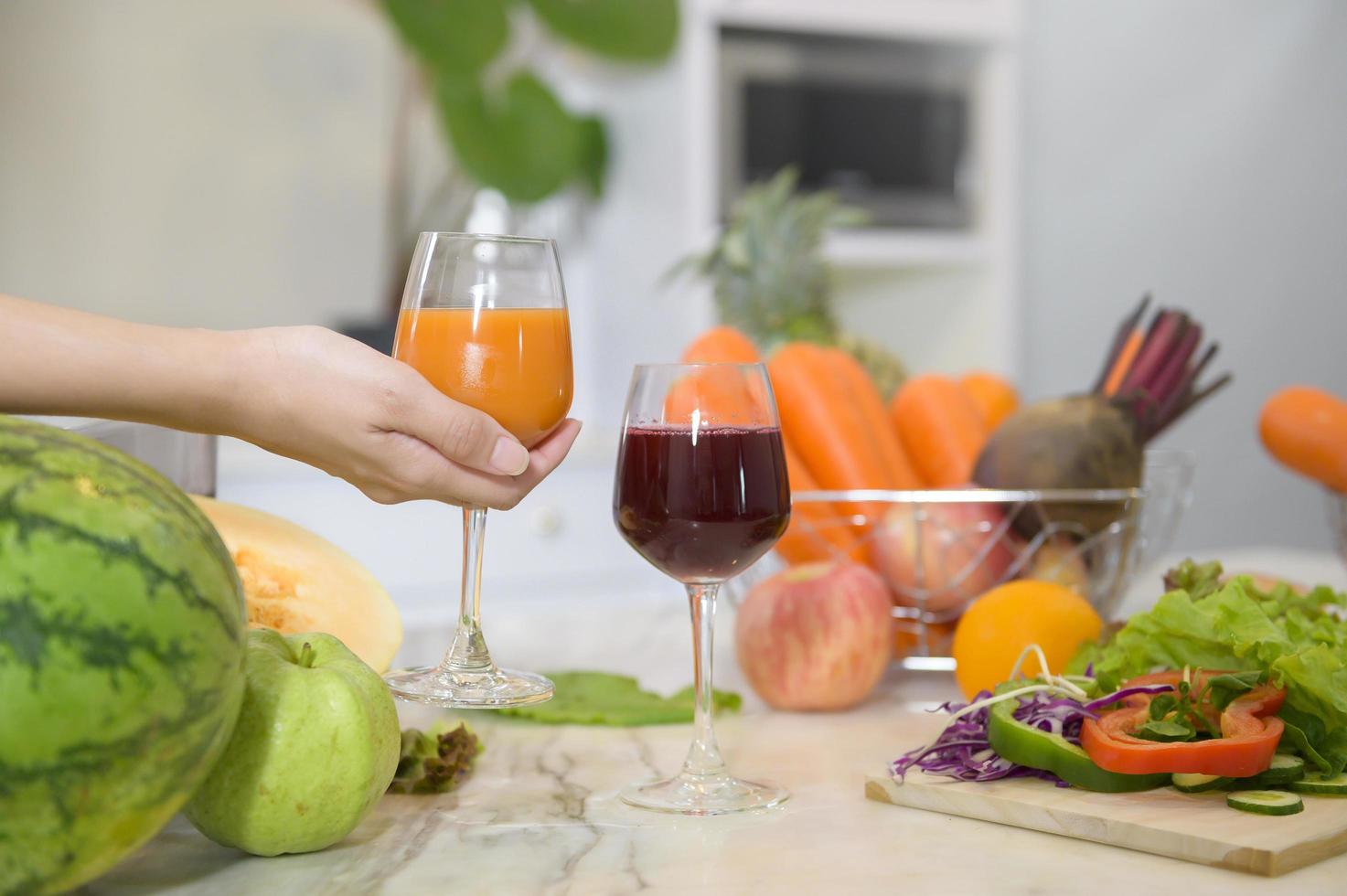 primo piano della mano che tiene un bicchiere di succo sano, mentre verdure e spremiagrumi sul tavolo in cucina, concetto di salute foto