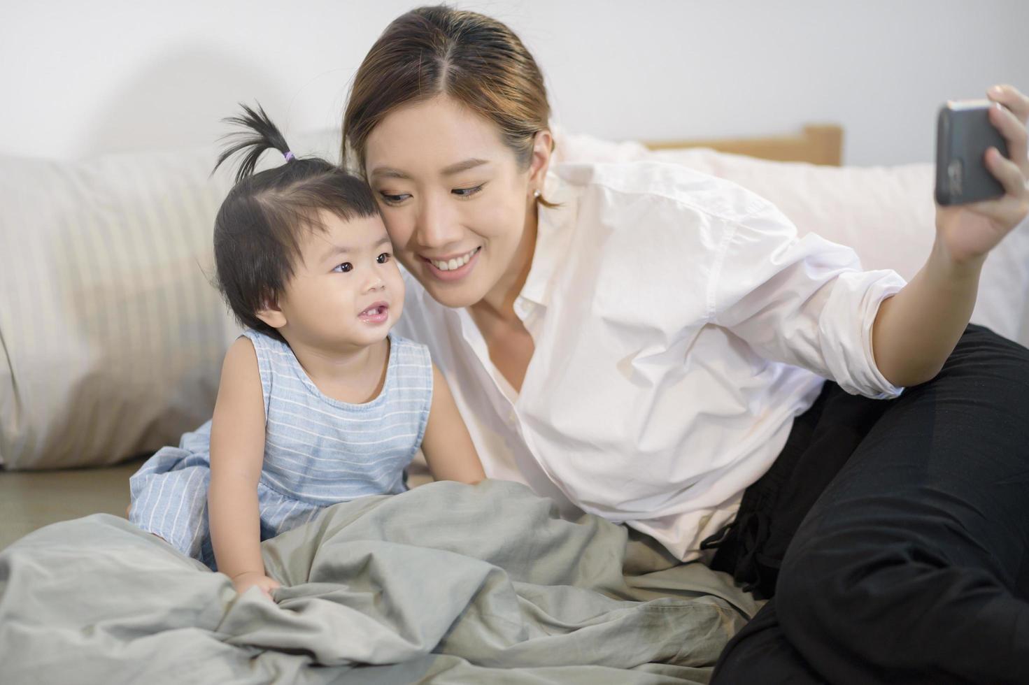 la madre asiatica e la sua bambina stanno facendo selfie o videochiamate al padre a letto, famiglia, sicurezza domestica, genitorialità, concetto tecnologico foto