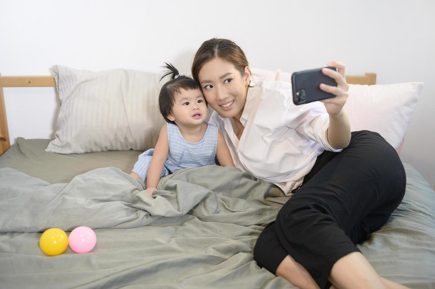 la madre asiatica e la sua bambina stanno facendo selfie o videochiamate al padre a letto, famiglia, sicurezza domestica, genitorialità, concetto tecnologico foto