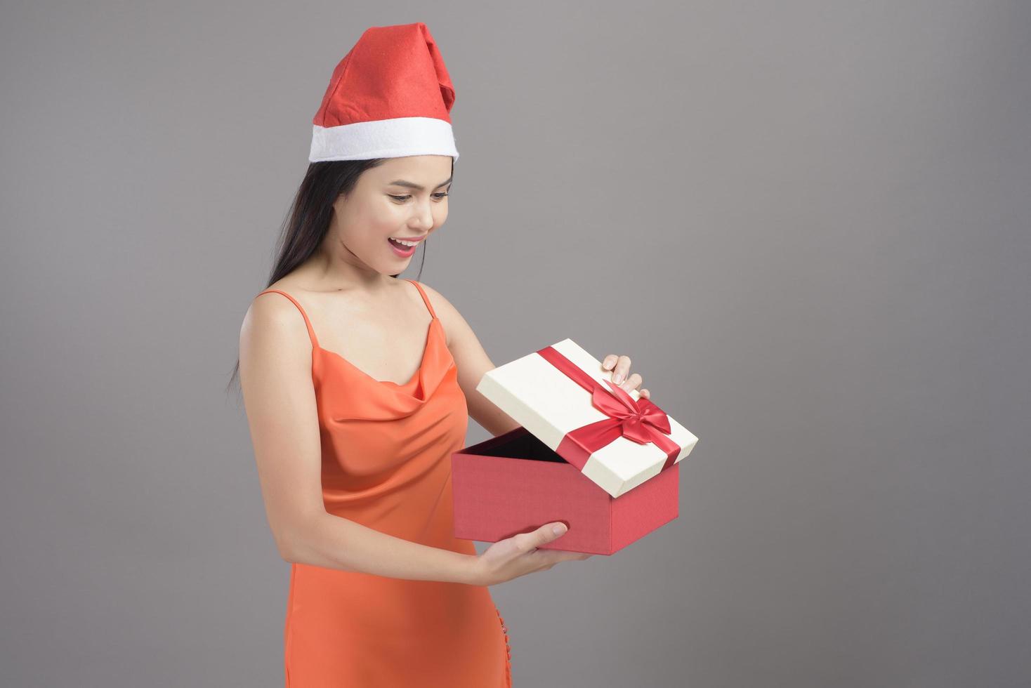 il ritratto di giovane donna sorridente che indossa il cappello rosso di Babbo Natale è in possesso di una confezione regalo isolata su sfondo grigio studio, concetto di natale e capodanno. foto