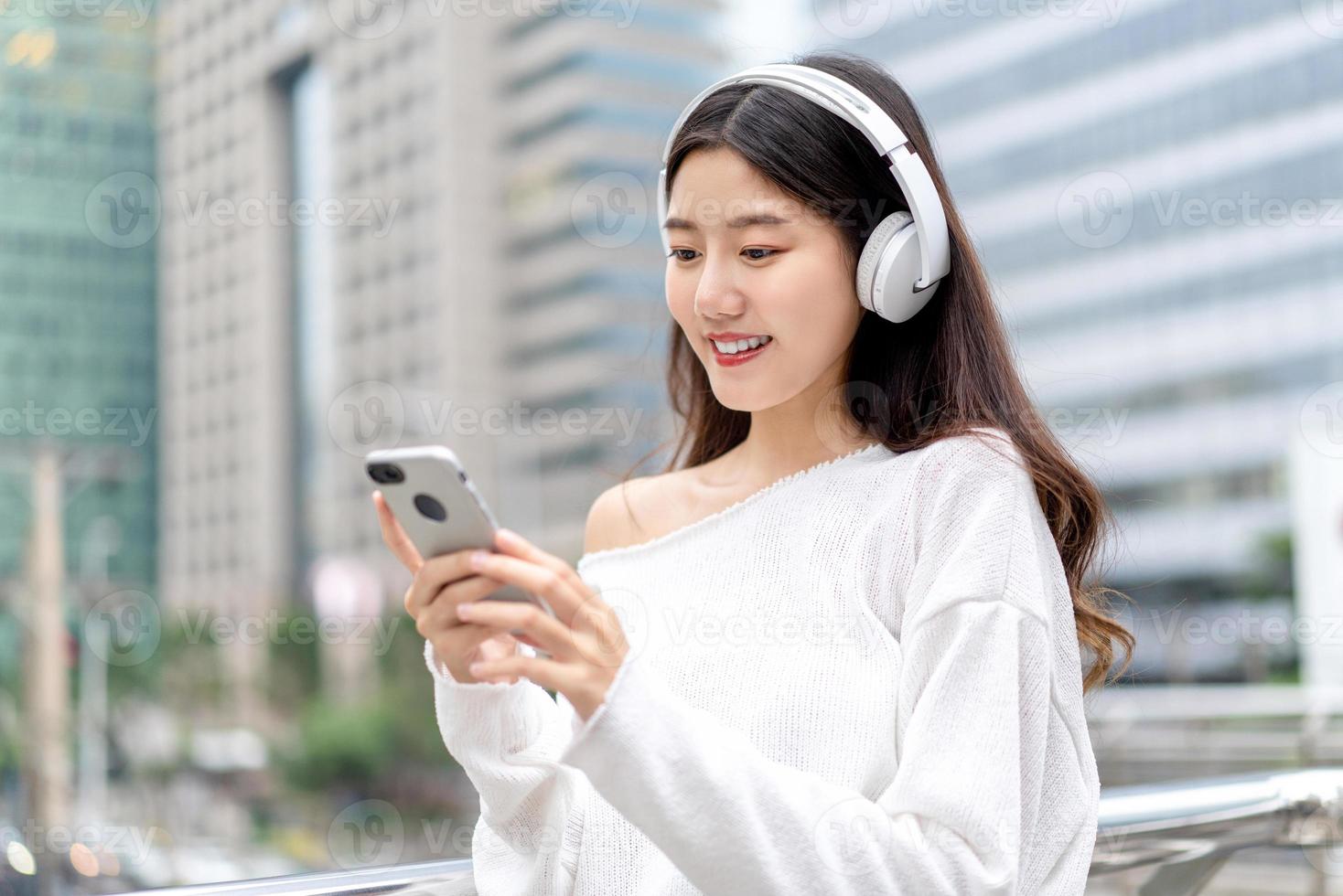 giovane ragazza asiatica che indossa le cuffie per ascoltare musica dal telefono cellulare contro il backrgound della costruzione della città foto