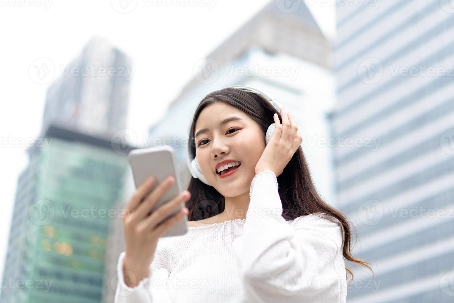donna asiatica sorridente felice che indossa le cuffie e guarda il telefono cellulare mentre si ascolta musica in streaming sullo sfondo dell'edificio della città foto