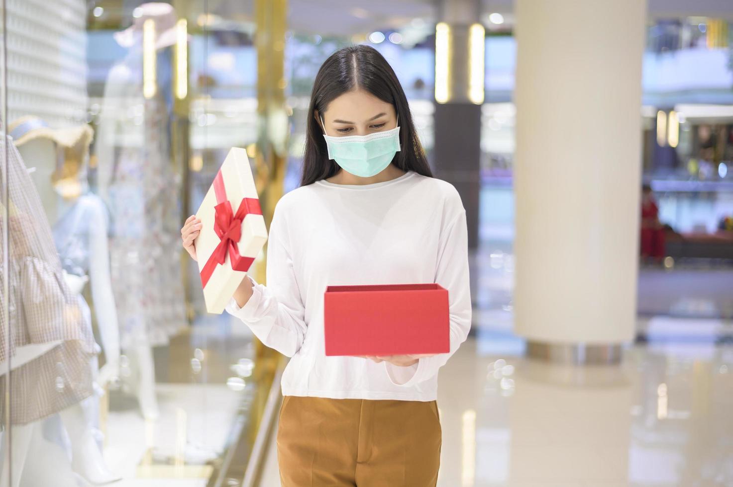 donna che indossa una maschera protettiva con in mano una confezione regalo nel centro commerciale, lo shopping sotto la pandemia covid-19, il ringraziamento e il concetto di natale. foto